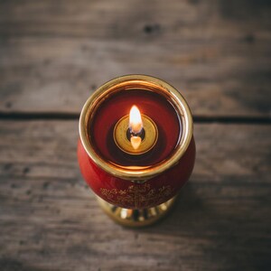 May include: A lit red candle in a gold-rimmed holder. The candle has a small flame and sits on a wooden surface. The candle holder has gold accents and decorative patterns.