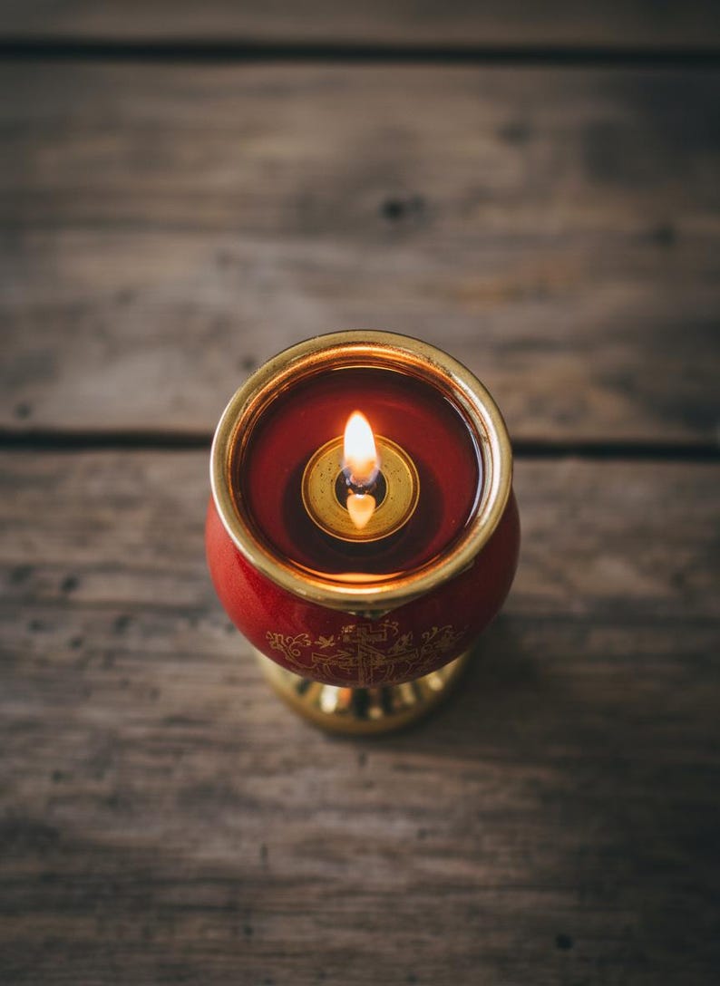 May include: A lit red candle in a gold-rimmed holder. The candle has a small flame and sits on a wooden surface. The candle holder has gold accents and decorative patterns.