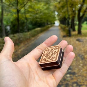 May include: A small, rectangular wooden box with a carved floral design on the lid. The box is held in a hand, showcasing its intricate details and the contrast between the light wood carving and the darker wood of the box.