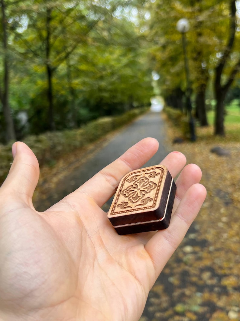 May include: A small, rectangular wooden box with a carved floral design on the lid. The box is held in a hand, showcasing its intricate details and the contrast between the light wood carving and the darker wood of the box.