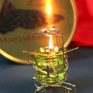 May include: A lit candle in a decorative gold-colored metal holder with a cross design. The candle is in a green glass cup, and a small metal dish sits above the flame. A gold-colored tin is in the background.