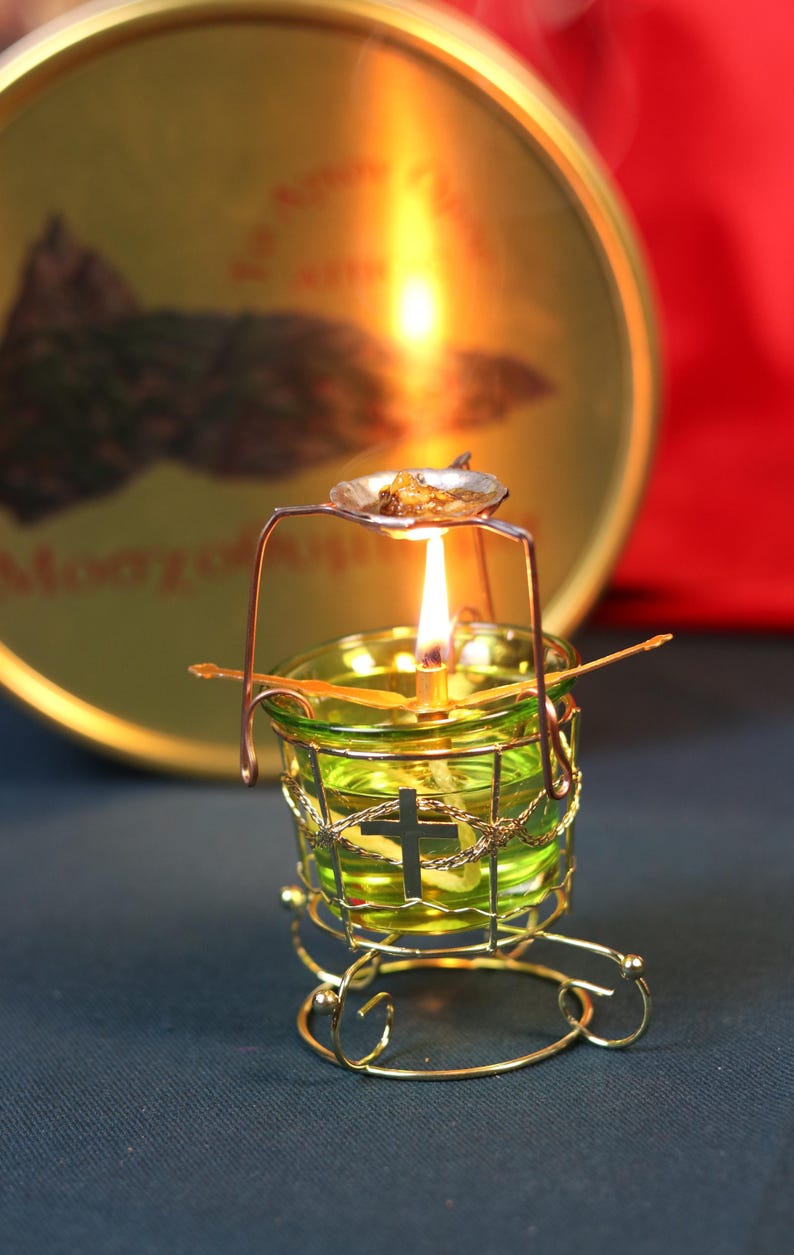 May include: A lit candle in a decorative gold-colored metal holder with a cross design. The candle is in a green glass cup, and a small metal dish sits above the flame. A gold-colored tin is in the background.