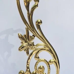 Puede incluir: Un soporte de metal dorado pulido con un brazo vertical en forma de S que presenta motivos florales decorativos y volutas. La estructura incluye un gancho curvado en la parte superior para colgar objetos y una base circular texturizada.