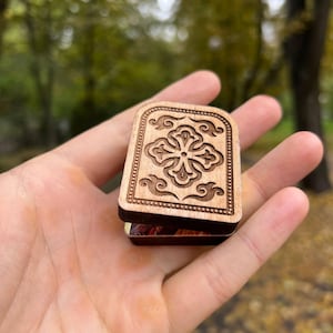 May include: A small, wooden box with a carved floral design on the lid. The box is rectangular with rounded edges and is held in an open hand. The background is blurred, with green and yellow foliage.