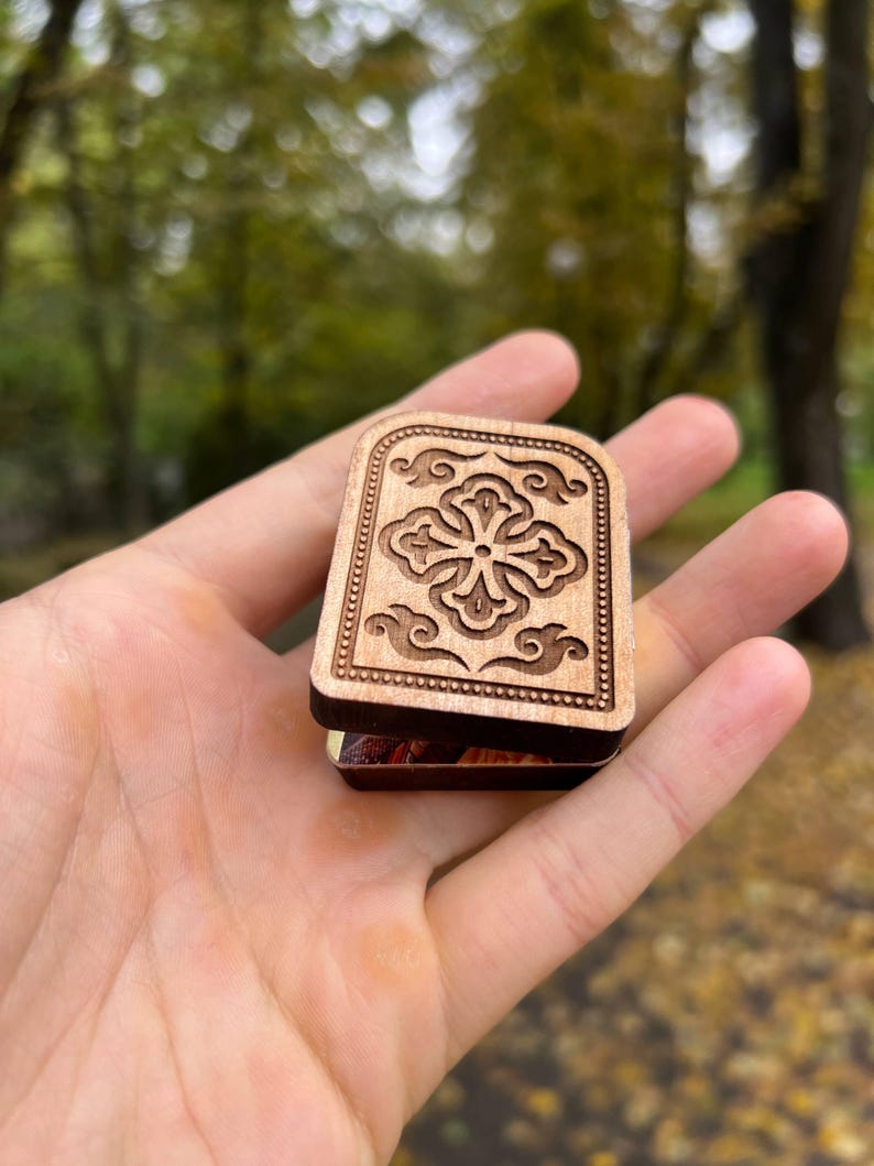May include: A small, wooden box with a carved floral design on the lid. The box is rectangular with rounded edges and is held in an open hand. The background is blurred, with green and yellow foliage.