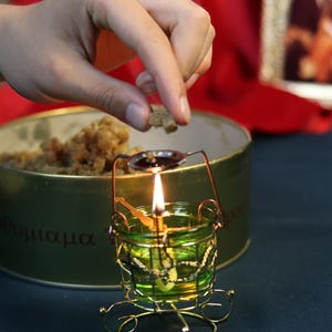 May include: A lit candle in a small, ornate green glass holder with a gold wire frame. A hand is placing a small piece of something into a spoon-shaped holder above the flame. A green tin with Greek text is in the background.