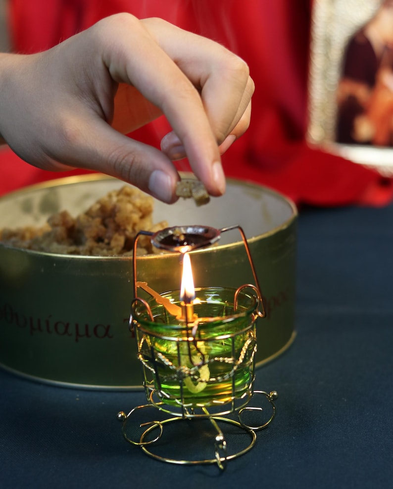 May include: A lit candle in a small, ornate green glass holder with a gold wire frame. A hand is placing a small piece of something into a spoon-shaped holder above the flame. A green tin with Greek text is in the background.