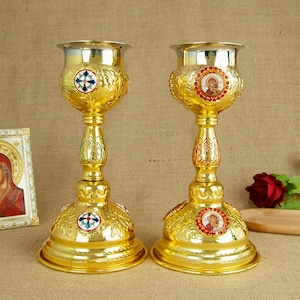 May include: Two ornate gold-colored chalices with silver rims. Each chalice features intricate embossed designs, decorative medallions, and a tall, slender stem. The chalices are likely used for religious ceremonies.