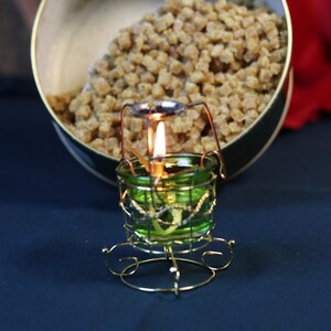May include: A lit green glass candle holder with a gold wire frame and a small flame. A container filled with small, brown, cube-shaped items is in the background.
