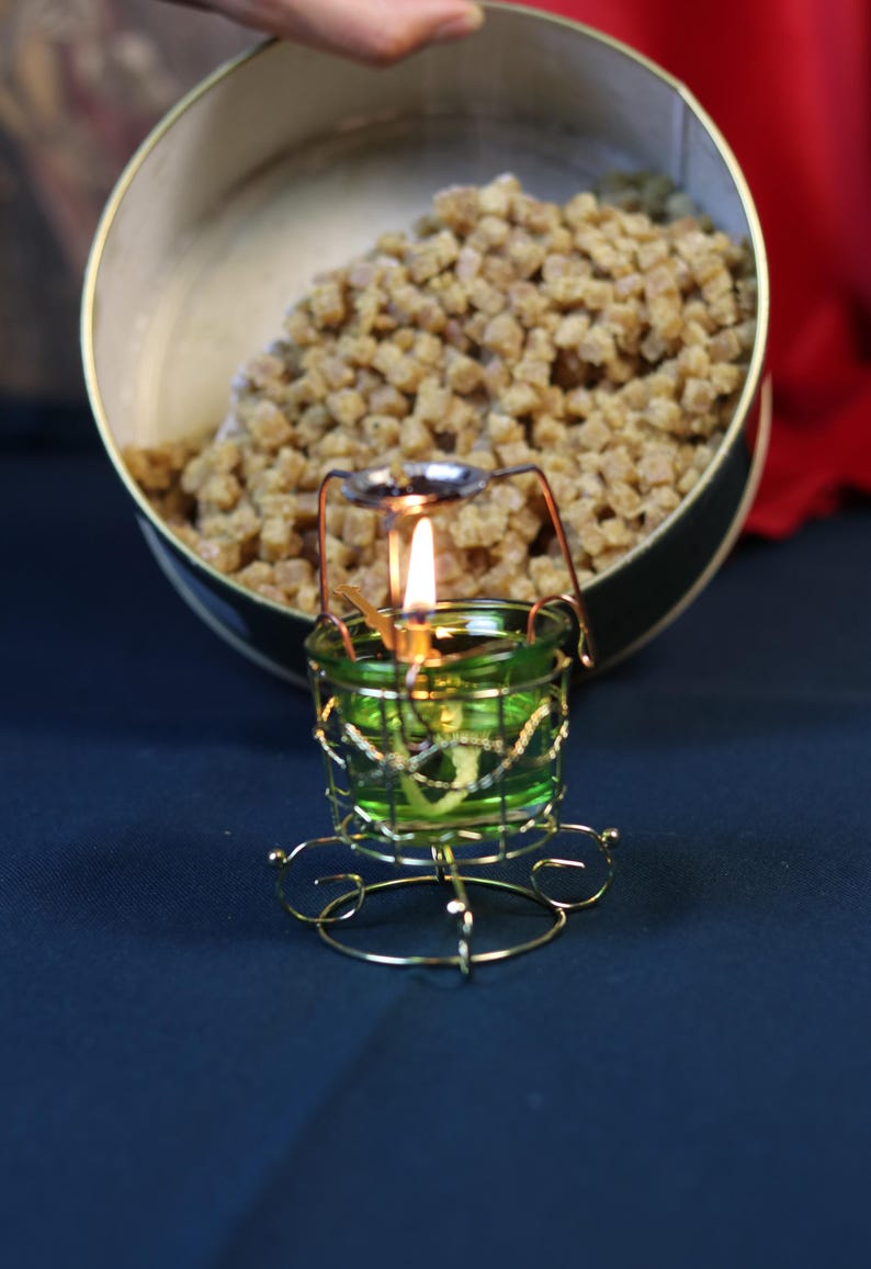 May include: A lit green glass candle holder with a gold wire frame and a small flame. A container filled with small, brown, cube-shaped items is in the background.