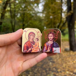 May include: Two small, gold-colored religious icons are held up. One depicts the Virgin Mary and child, the other Jesus Christ. The icons have detailed illustrations with red, orange, and gold tones. The background is blurred with green and brown.