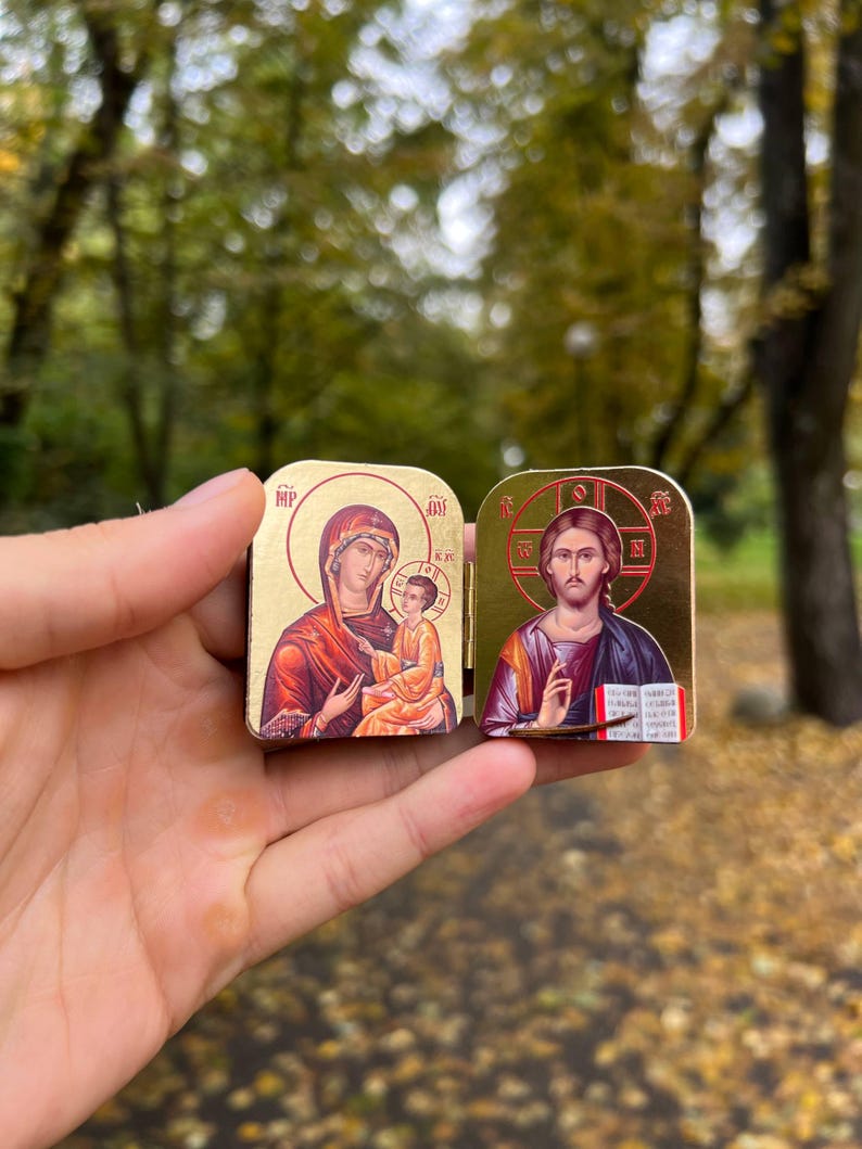 May include: Two small, gold-colored religious icons are held up. One depicts the Virgin Mary and child, the other Jesus Christ. The icons have detailed illustrations with red, orange, and gold tones. The background is blurred with green and brown.
