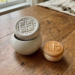 May include: Two small, round, ceramic containers with lids. One is a light beige color, and the other is a light brown color. Each lid features a cross design. The containers are on a wooden surface.