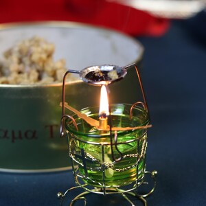 May include: A lit candle in a green glass holder with a gold wire cage and base. A small spoon with dark contents sits above the flame. The background includes a blurred container and red fabric.