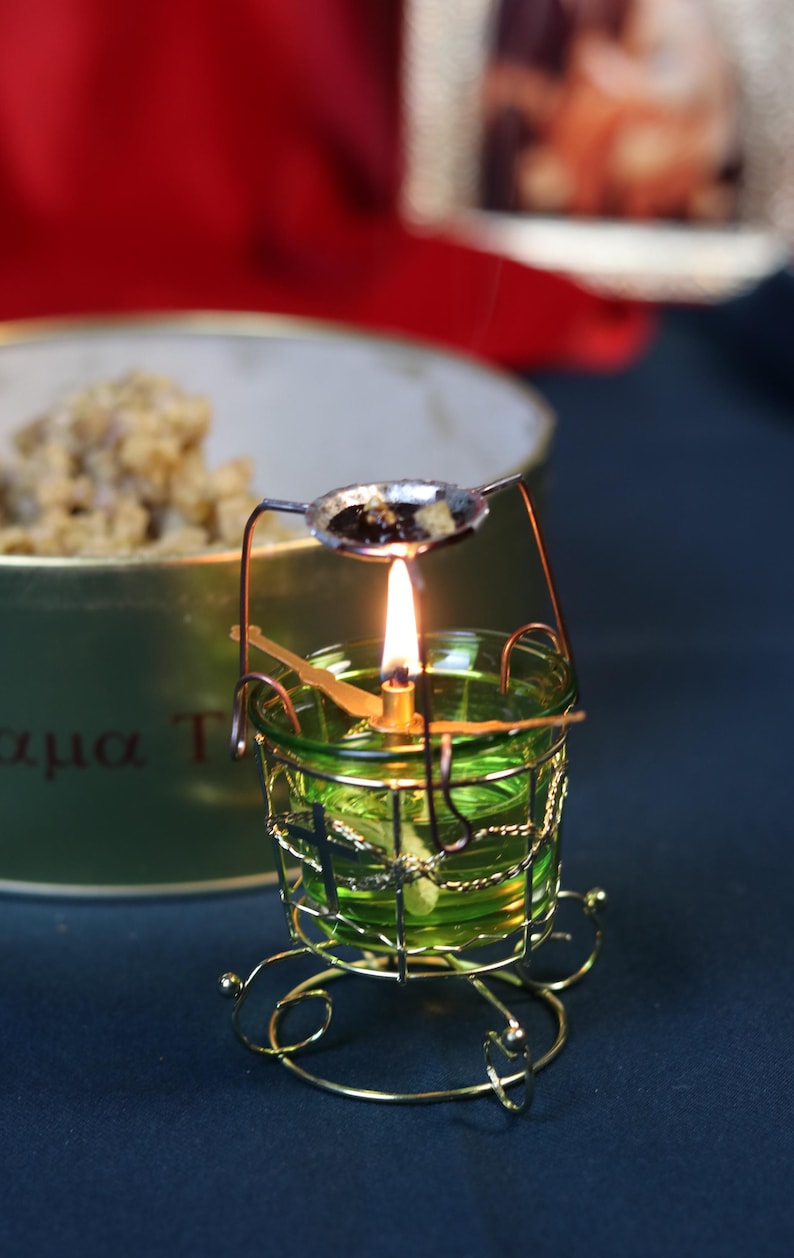 May include: A lit candle in a green glass holder with a gold wire cage and base. A small spoon with dark contents sits above the flame. The background includes a blurred container and red fabric.