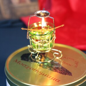May include: A lit candle in a small, ornate, gold-colored metal holder with a green glass cup. The holder sits atop a round, gold-colored tin with red text. The flame is bright against a blurred background.