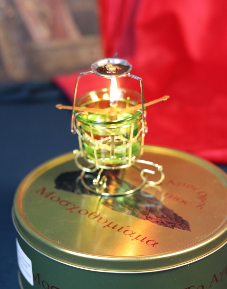 May include: A lit candle in a small, ornate, gold-colored metal holder with a green glass cup. The holder sits atop a round, gold-colored tin with red text. The flame is bright against a blurred background.