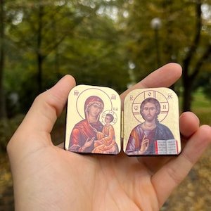 May include: Two small, gold-colored religious icons are held in a hand. One depicts a woman and child, the other a man. The icons have a rectangular shape with rounded tops and feature detailed illustrations with religious symbols.