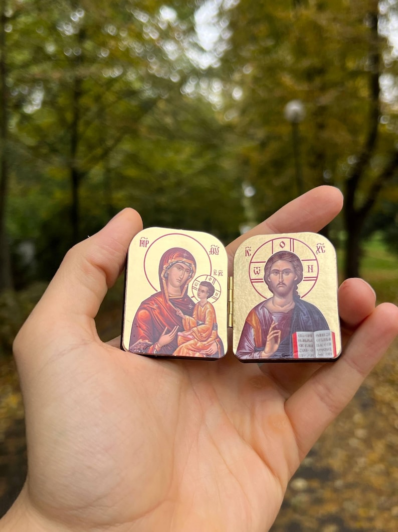May include: Two small, gold-colored religious icons are held in a hand. One depicts a woman and child, the other a man. The icons have a rectangular shape with rounded tops and feature detailed illustrations with religious symbols.