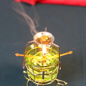 May include: A lit candle in a green glass holder with a gold-colored metal frame. A small dish with a substance is above the flame, emitting smoke. The candle holder has a cross design.