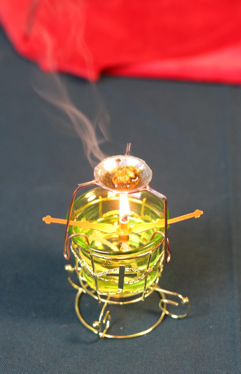 May include: A lit candle in a green glass holder with a gold-colored metal frame. A small dish with a substance is above the flame, emitting smoke. The candle holder has a cross design.