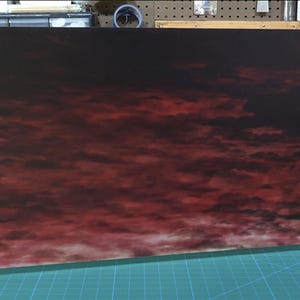 Könnte beinhalten: Ein rechteckiges Panel mit einer dramatischen Himmelsansicht in tiefroten und schwarzen Farbtönen, die an einen Sonnenuntergang oder einen Sturm erinnern. Das Panel wird von zwei kleinen weißen Ständern getragen, die auf einer grünen Rasteroberfläche stehen.