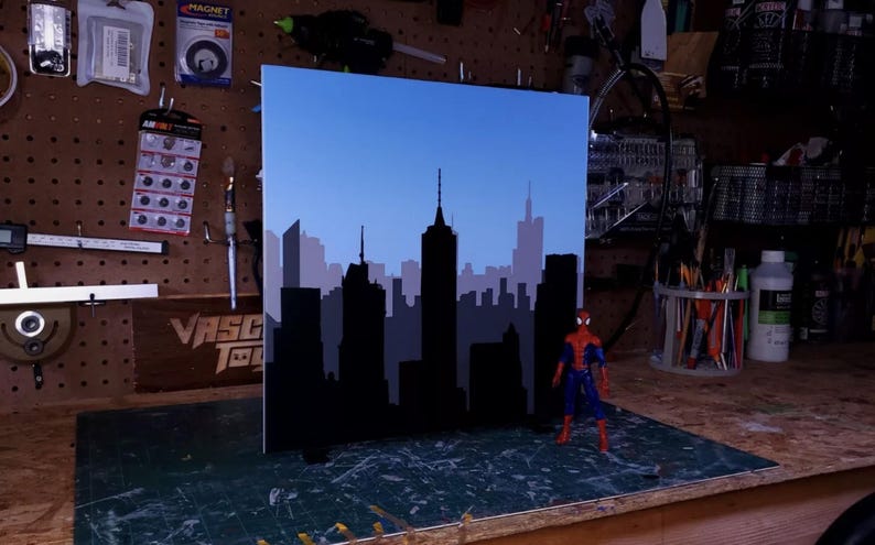 May include: A black and blue cityscape backdrop with a blue sky, perfect for use as a photography prop or diorama. The backdrop is made of cardboard and features a silhouette of a city skyline.