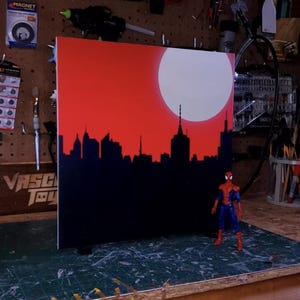 May include: A black and red cityscape painting with a white moon and a red background. A Spiderman action figure stands in front of the painting.