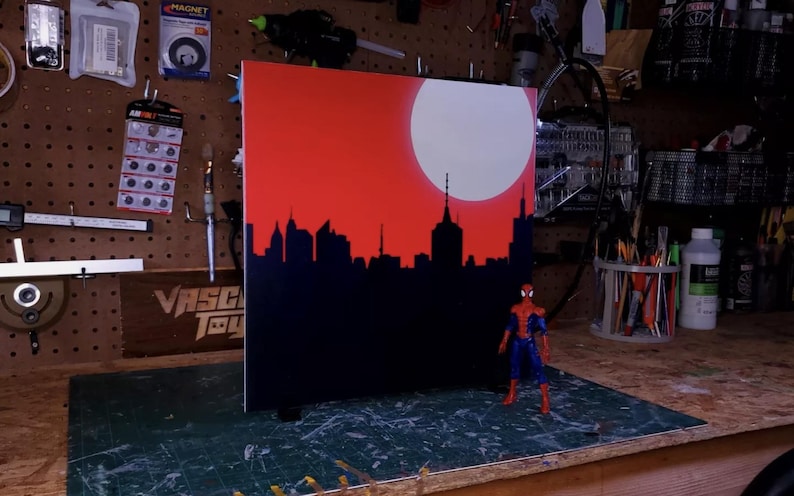 May include: A black and red cityscape painting with a white moon and a red background. A Spiderman action figure stands in front of the painting.