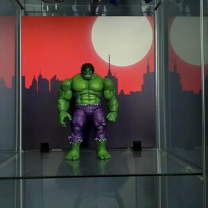 May include: A green Hulk action figure with purple pants standing on a glass shelf in front of a red and white cityscape backdrop.