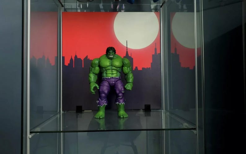 May include: A green Hulk action figure with purple pants standing on a glass shelf in front of a red and white cityscape backdrop.