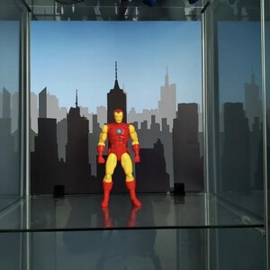 May include: A red and yellow action figure of Iron Man standing in front of a black cityscape backdrop. The figure is posed with its arms outstretched and its head tilted slightly to the side. The figure is standing on a glass shelf.