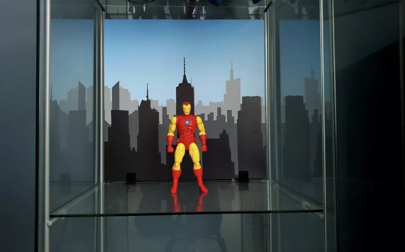May include: A red and yellow action figure of Iron Man standing in front of a black cityscape backdrop. The figure is posed with its arms outstretched and its head tilted slightly to the side. The figure is standing on a glass shelf.