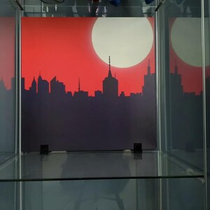 May include: A glass display case with a red and white cityscape print on the back wall. The cityscape is a silhouette of buildings against a red background with a white sun.