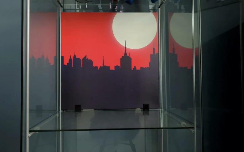 May include: A glass display case with a red and white cityscape print on the back wall. The cityscape is a silhouette of buildings against a red background with a white sun.