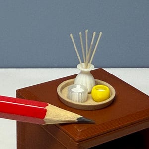 May include: A miniature set featuring a small white vase with thin sticks, a translucent white cylindrical container, and a small yellow bowl, all arranged on a circular tan tray. A red pencil is positioned in the foreground for scale.