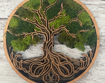 May include: A round wooden wall art piece featuring a layered tree design. The tree's branches and roots are intricately cut from wood, with green moss filling the tree's crown. The artwork is set against a dark brown background and framed by a light brown border.