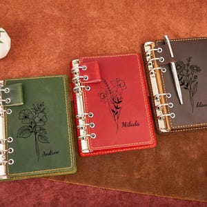 Puede incluir: Tres diarios de cuero en verde, rojo y marrón, cada uno con una encuadernación plateada. Los diarios presentan arte lineal floral y nombres personalizados: "Andrew", "Milada" y "blanch". Un bolígrafo plateado descansa sobre el diario marrón.