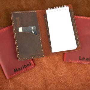 Puede incluir: Juego de cuaderno y tarjetero de cuero en marrón y rojo. El cuaderno abierto tiene un pequeño bloc de notas y una ranura para tarjetas. Se ven dos tarjeteros rojos, uno con el nombre "Maribel" y otro con "Leah".
