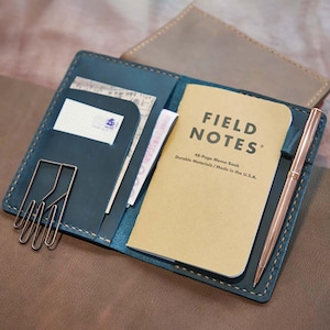 May include: Open teal leather notebook cover with a Field Notes memo book, a rose gold pen, and a paper clip. The memo book is tan with the words "FIELD NOTES" printed on the cover. The cover has pockets for cards and other small items.