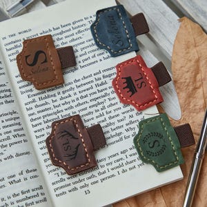 Peut inclure: Cinq porte-stylos en cuir bleu, marron, rouge, marron foncé et vert, chacun doté d'une bande élastique marron et de gravures personnalisées incluant le nom Selena, les initiales SL, une couronne, des bois et une couronne de laurier.