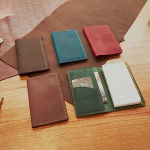May include: A selection of handmade leather notebooks and wallets in various colours, including brown, teal, and red. One wallet is open, revealing a notepad, credit card, and other items. The notebooks and wallets are arranged on a wooden surface.