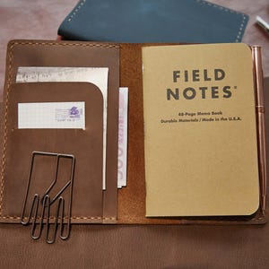 May include: Open brown leather notebook with a Field Notes memo book, a pen, and a metal paper clip. The notebook has pockets holding a postcard and currency. The Field Notes book is made of durable materials and made in the U.S.A.