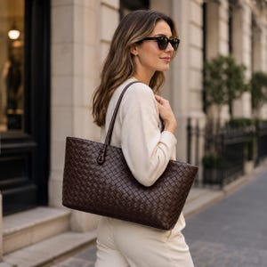 Puede incluir: Un bolso tote de cuero marrón oscuro que presenta un intrincado patrón trenzado en toda la superficie exterior. El bolso tiene dos correas de hombro finas de cuero a juego, unidas con detalles anudados en el borde superior del cuerpo rectangular.