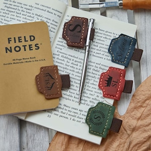 Op de afbeelding: Een verzameling gepersonaliseerde leren penhouders en een Field Notes notitieboek. De penhouders zijn er in verschillende kleuren, elk met een uniek ontwerp en initialen of een naam. Het notitieboek is bruin met de tekst "FIELD NOTES" erop.