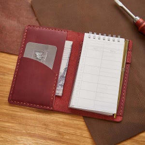 Puede incluir: Una funda de cuaderno de cuero burdeos con un bloc de notas, un bolígrafo y ranuras para tarjetas. El bloc de notas está encuadernado en espiral y tiene un bolígrafo dorado insertado. Una tarjeta metálica y una foto de la Torre Eiffel están en los bolsillos.