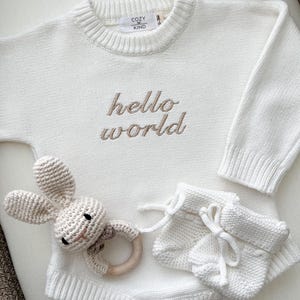 May include: A cream-colored baby outfit set, including a sweater with "hello world" embroidery, a romper, and matching booties. A crocheted bunny rattle with a wooden ring is also included. The sweater has the text "COZY KIND" on the label.