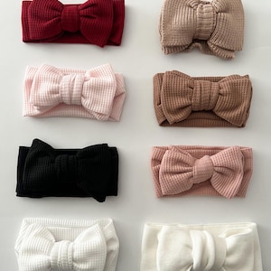 Puede incluir: Un surtido de ocho diademas para bebés en varios colores, incluyendo burdeos, beige, rosa, marrón, negro y blanco. Cada diadema presenta un lazo grande en el centro. Las diademas están hechas de una tela texturizada.