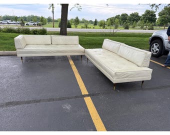 May include: Two-piece cream-colored sofa set with a mid-century modern design. The set includes a sofa with a single armrest and a chaise lounge. Both pieces have wooden legs and a tufted seat.