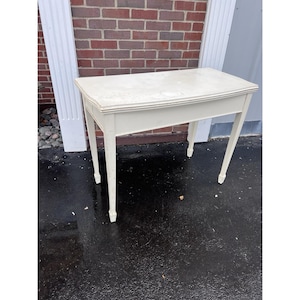 May include: A small, off-white painted table with a rectangular top and four slender, tapered legs. The table has a simple, classic design, suitable for various uses. The paint shows signs of wear, adding to its vintage appeal.
